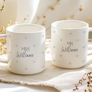 May include: Two white ceramic mugs with handles. The mug on the left has pink hearts and the text "MRS Williams". The mug on the right has blue hearts and the text "MR Williams".