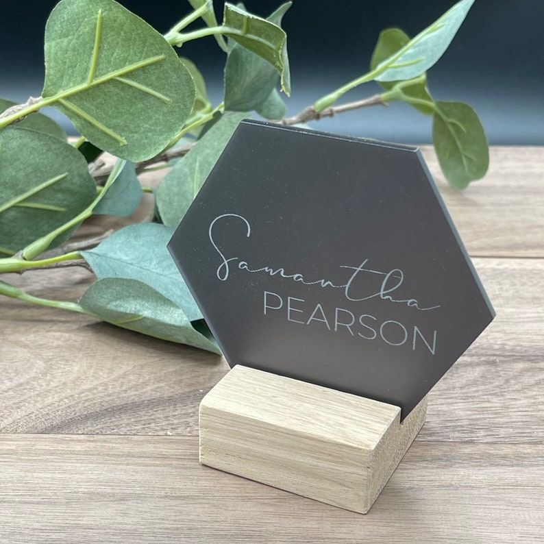 Pu&ograve; includere: Un biglietto da visita esagonale grigio scuro con il nome "Samantha Pearson" in corsivo. Il biglietto &egrave; tenuto in un blocco di legno chiaro. Foglie verdi sullo sfondo.