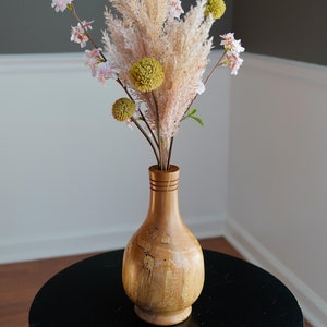 May include: A wooden vase with a light brown finish holds a floral arrangement. The arrangement includes pampas grass, pink blossoms, and green, spherical flowers. The vase sits on a black table.