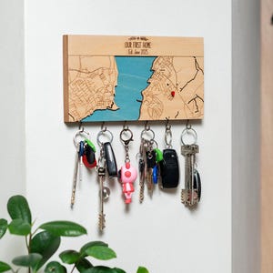 May include: Wooden key holder with a map design, blue water, and a red pin. The text "OUR FIRST HOME Est. June 2025" is printed on the top. Several keys hang from the bottom. The key holder is mounted on a white wall.