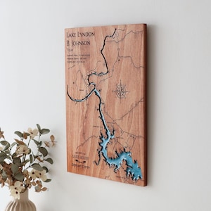 Könnte beinhalten: Wandkunst aus Holz, die eine Karte des Lake Lyndon B. Johnson in Texas zeigt. Der See ist blau dargestellt, das umliegende Land in einem natürlichen Holzton. Der Text enthält den Namen des Sees, den Standort und weitere Details. Eine Windrose ist ebenfalls abgebildet.