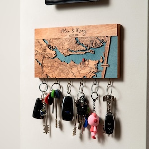 May include: Wooden key holder with a detailed map design, featuring a blue water element and a red marker. The key holder is personalized with the names "Alex & Mary" and the phrase "Welcome Home". Several keys hang from the bottom.
