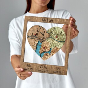 Framed wooden wall art featuring a heart-shaped puzzle of layered location maps with red pins, personalized with couple names on top and dates on the bottom.