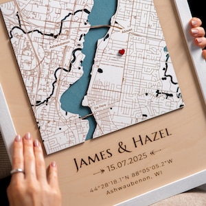 May include: A framed map with a white border, featuring a detailed street layout in brown and a blue water feature. A red heart marks a location. The text "JAMES & HAZEL" and a date are engraved below, along with coordinates and the city name.