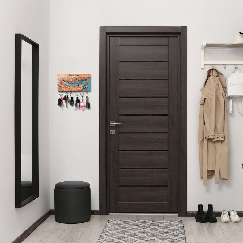 Full entryway scene with a custom map key holder mounted on the wall near a dark wood door, mirror, bench, and coat rack. The piece reads as functional minimalist wall decor designed for organized modern living.