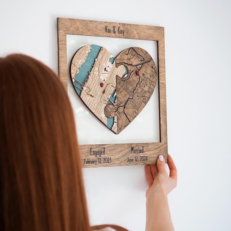 A woman hangs a personalized wooden heart map on the wall, showing the piece as custom wall art for a couple’s engagement and wedding story. The image reinforces the product as a meaningful display keepsake that connects two locations in one heart.