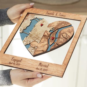 Hands hold a personalized wooden heart map wall art piece showing two connected locations with custom names and engaged and married dates. This close view highlights the layered craftsmanship and makes the custom engagement and wedding gift feel personal and gift-ready.