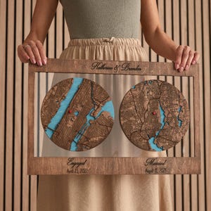 May include: A rectangular wooden frame containing two circular laser-cut maps with blue water details and red heart markers. The top is engraved with "Katherine & Brandon" and the bottom with "Engaged April 23, 2022" and "Married August 13, 2023".