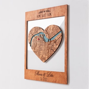 Può includere: Una cornice rettangolare in legno con una mappa a forma di cuore tagliata al laser, un inserto a fiume blu e una piccola icona di casa rossa. Il bordo superiore mostra HOME SWEET HOME, quello inferiore mostra Maria & Peter e Est. 2022.