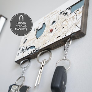 May include: A wooden key holder shaped like a map of New Orleans, Louisiana. The map is white with brown lines and a blue body of water. The key holder has three hooks for hanging keys. The key holder has a red heart marking the location of the French Quarter. The key holder has a dark brown wood frame.