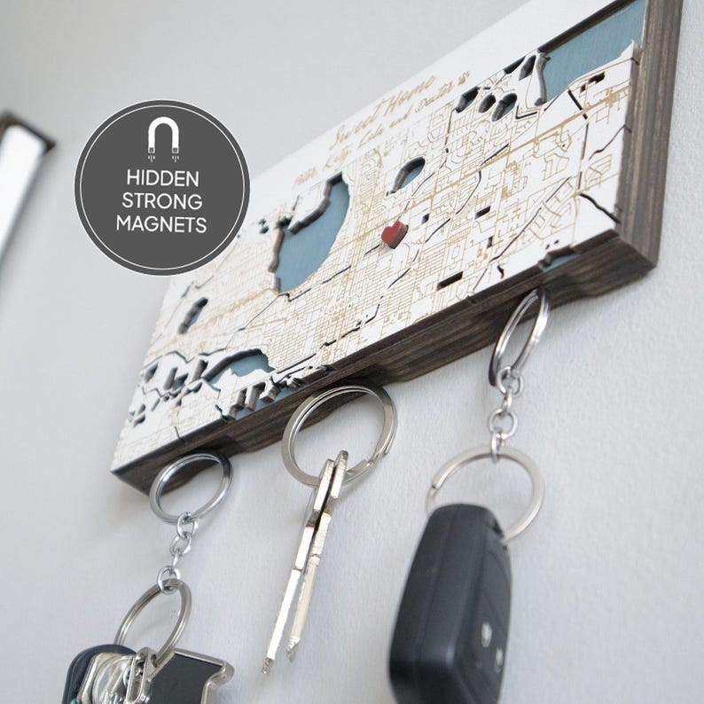 May include: A wooden key holder shaped like a map of New Orleans, Louisiana. The map is white with brown lines and a blue body of water. The key holder has three hooks for hanging keys. The key holder has a red heart marking the location of the French Quarter. The key holder has a dark brown wood frame.