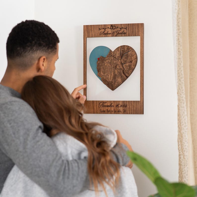 May include: A wooden heart-shaped map with a blue and brown color scheme. The map is framed in a dark wood frame and includes the text "Andrew & Jocelyn" and "November 16, 2023,  Coroner Park in La Jolla, CA".