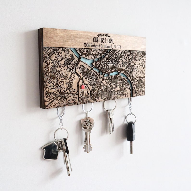 May include: Wooden key holder with a detailed map design, featuring the text "OUR FIRST HOME" and address details. The map has a light blue river and a red pin. Several keys hang from the bottom.