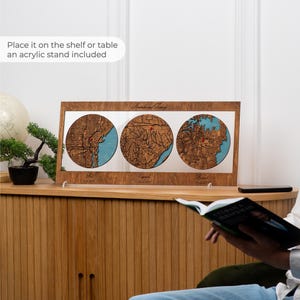 May include: A rectangular wooden frame containing three circular laser-cut maps with blue water details and red pin markers. The frame features engraved text and is supported by a clear acrylic stand, displaying geographic locations.