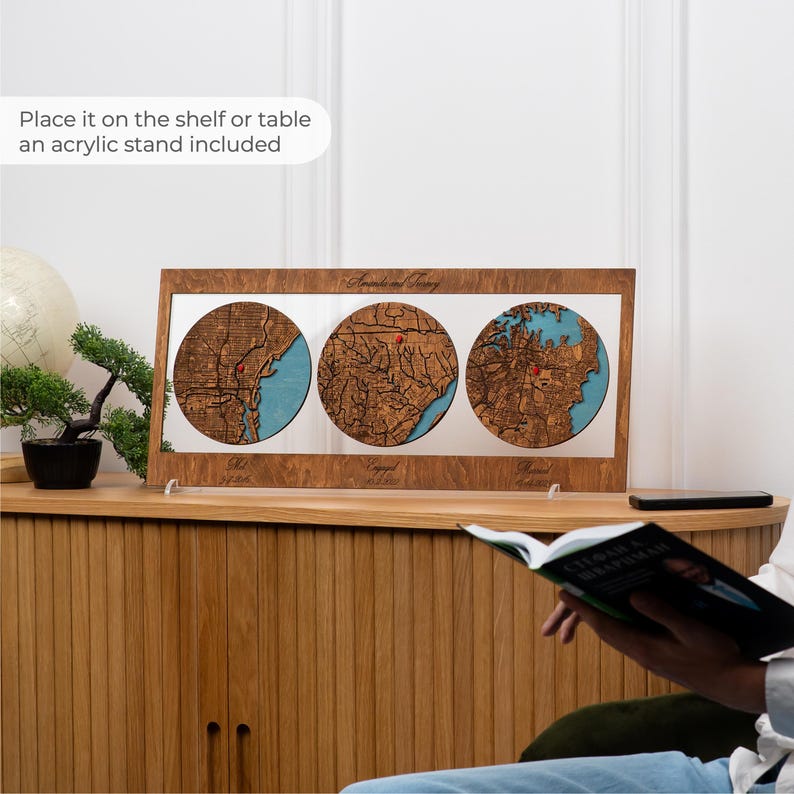 May include: A rectangular wooden frame containing three circular laser-cut maps with blue water details and red pin markers. The frame features engraved text and is supported by a clear acrylic stand, displaying geographic locations.