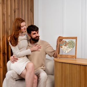 A seated couple poses with the framed heart puzzle map placed on a nearby cabinet, creating a warm romantic home scene. This image shows the personalized piece as a wedding keepsake gift made to honor a couple’s journey together.
