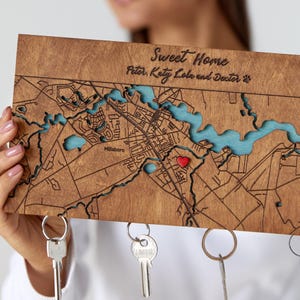 May include: Wooden key holder with a map design, featuring a town layout with blue water features and a red heart marker. The text "Sweet Home" and names are engraved at the top. Includes key rings for hanging keys.