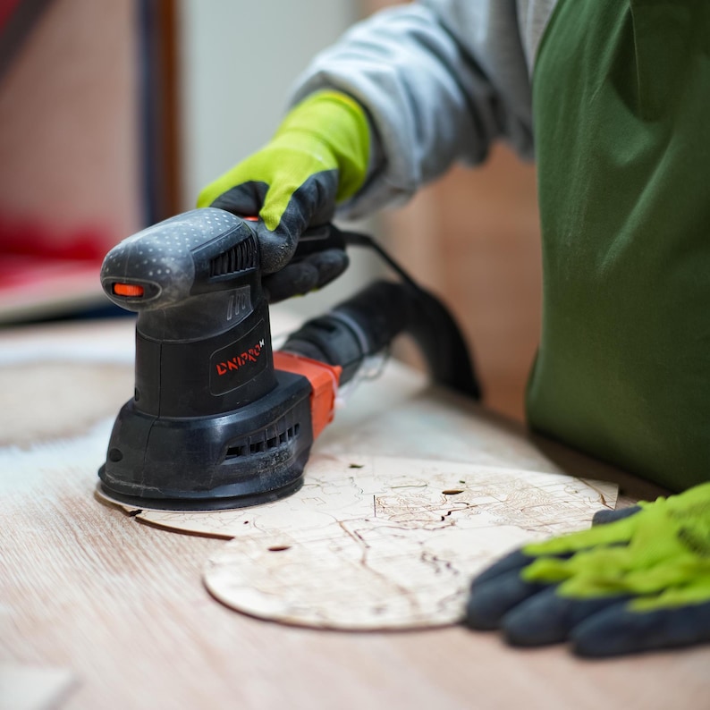 Close-up of a sanding tool finishing a wooden heart puzzle map piece on a workbench. This photo highlights the handcrafted production process used to create the personalized wedding keepsake.
