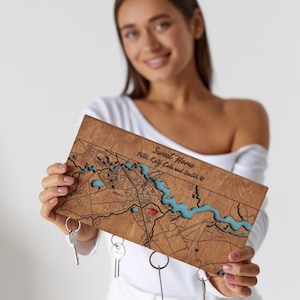 A woman holds a personalized wooden map key holder with keys attached below by hidden magnets. The photo shows the product scale, warm wood finish, and custom home location design.