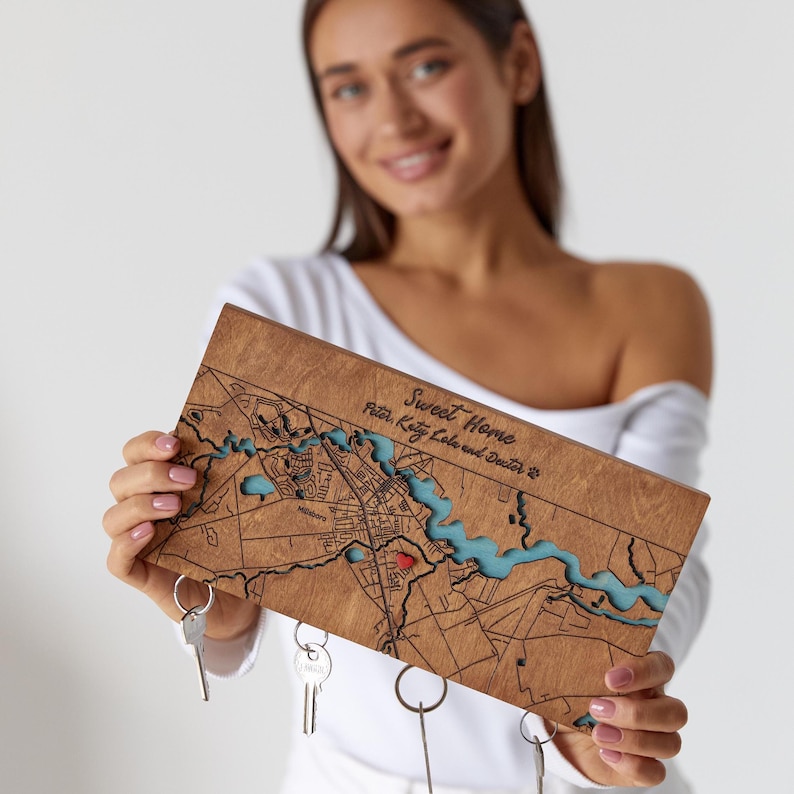 A woman holds a personalized wooden map key holder with keys attached below by hidden magnets. The photo shows the product scale, warm wood finish, and custom home location design.