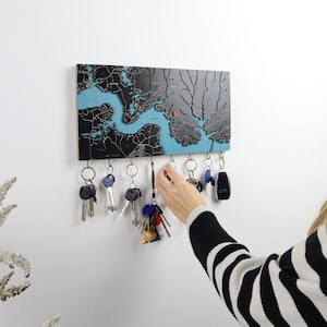 A person reaches for keys on a dark wood map key rack mounted on a white wall. This custom entryway organizer shows how the wall key holder works for everyday use in a modern home.