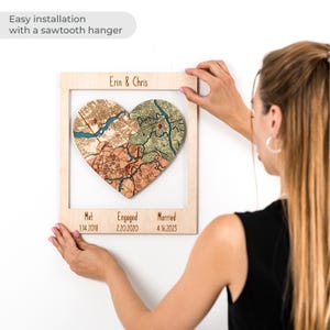 A woman hangs the framed heart puzzle map on the wall, with a note about easy installation using a sawtooth hanger. This image shows the personalized wedding keepsake as ready-to-hang wall decor for a couple’s home.