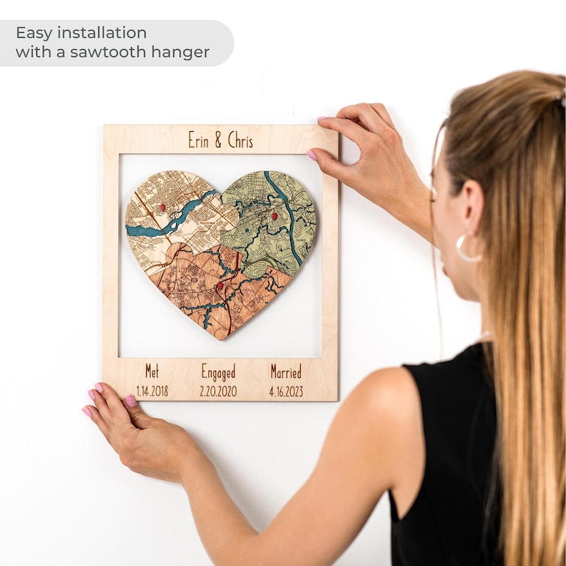 A woman hangs the framed heart puzzle map on the wall, with a note about easy installation using a sawtooth hanger. This image shows the personalized wedding keepsake as ready-to-hang wall decor for a couple’s home.