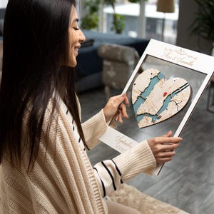 May include: A white wooden frame with a heart-shaped cutout. The cutout is filled with a map of a city in a blue and white color scheme. The text "September 4th, 2022" is written on the frame. The frame is being held by a person.