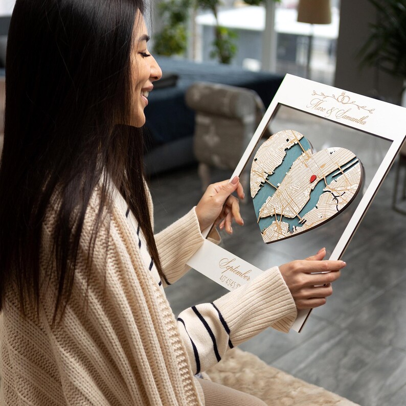 May include: A white wooden frame with a heart-shaped cutout. The cutout is filled with a map of a city in a blue and white color scheme. The text "September 4th, 2022" is written on the frame. The frame is being held by a person.