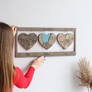 Könnte beinhalten: Ein rechteckiges Wandbild aus Holz mit drei herzförmigen Ausschnitten, die eingravierte Kartendetails zeigen. Der Rahmen trägt oben den Text "Andrea & Jordan" und unten "Hello", "Will you?" sowie "I do". Jedes Herz enthält eine kleine rote Stecknadel.