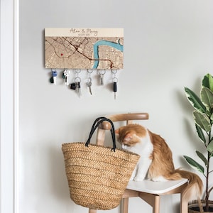May include: A wooden key holder with a map design, personalized with the names "Alex & Mary" and "Welcome Home". The key holder has several keys hanging from it. A woven straw tote bag and a cat are in the foreground.