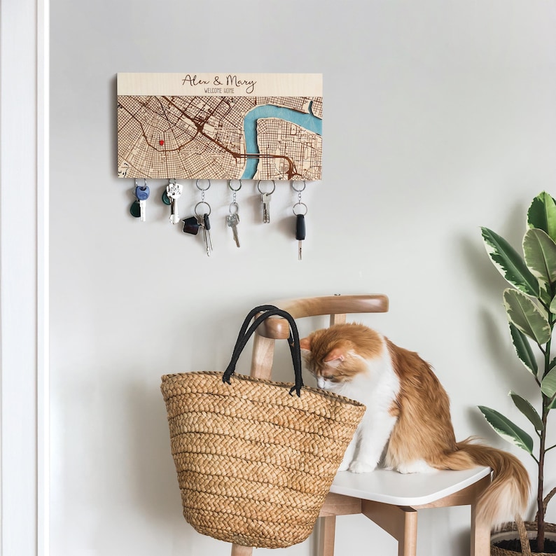 May include: A wooden key holder with a map design, personalized with the names "Alex & Mary" and "Welcome Home". The key holder has several keys hanging from it. A woven straw tote bag and a cat are in the foreground.