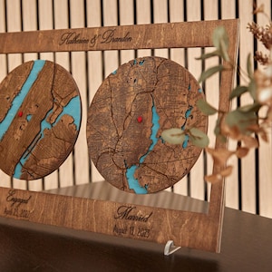 May include: A rectangular wooden frame containing two circular laser-cut maps with blue water details and red pin markers. The frame features the engraved text Katherine & Brandon, Engaged April 23, 2022, and Married August 13, 2023.