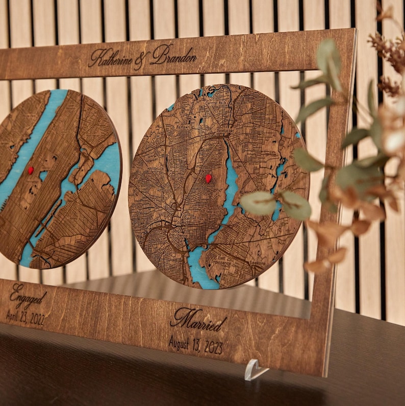 May include: A rectangular wooden frame containing two circular laser-cut maps with blue water details and red pin markers. The frame features the engraved text Katherine & Brandon, Engaged April 23, 2022, and Married August 13, 2023.