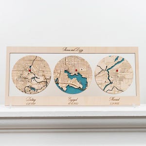 May include: A rectangular light wood frame containing three circular laser-cut wooden maps with teal water details. Each map features a small red pin. The text reads Thomas and Lizzy, Dating 4.27.2021, Engaged 12.15.2023, and Married 5.31.2025.