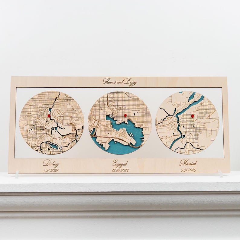 May include: A rectangular light wood frame containing three circular laser-cut wooden maps with teal water details. Each map features a small red pin. The text reads Thomas and Lizzy, Dating 4.27.2021, Engaged 12.15.2023, and Married 5.31.2025.