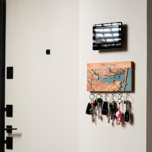 A personalized map key rack with hooks is mounted in a modern entryway beneath a wall panel, holding everyday keys. The lifestyle scene shows how the custom wall organizer adds order without cluttering the space.