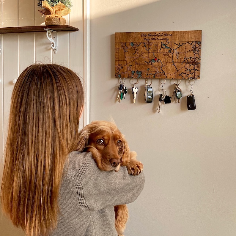May include: A wooden key holder with a map design and the text "Our Wonderful Home" engraved on it. The key holder has five hooks for hanging keys. A brown dog is sitting on the shoulder of a person.