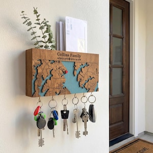 May include: A wooden key holder with a map design, featuring a blue resin inlay and a red dot. The key holder has a shelf for mail and a row of hooks for keys. The text "Collins Family" and coordinates are visible.