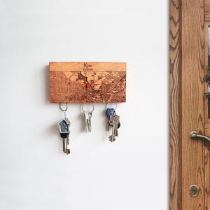 May include: Wooden key holder with a map of a city and the name "Kim" engraved on it. The key holder has three hooks for keys. There are three keys hanging on the hooks.