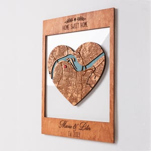 A framed heart-shaped map is shown from an angled view to highlight the depth and layered wood construction. This personalized wall art makes a thoughtful housewarming gift for a couple moving into a first home.