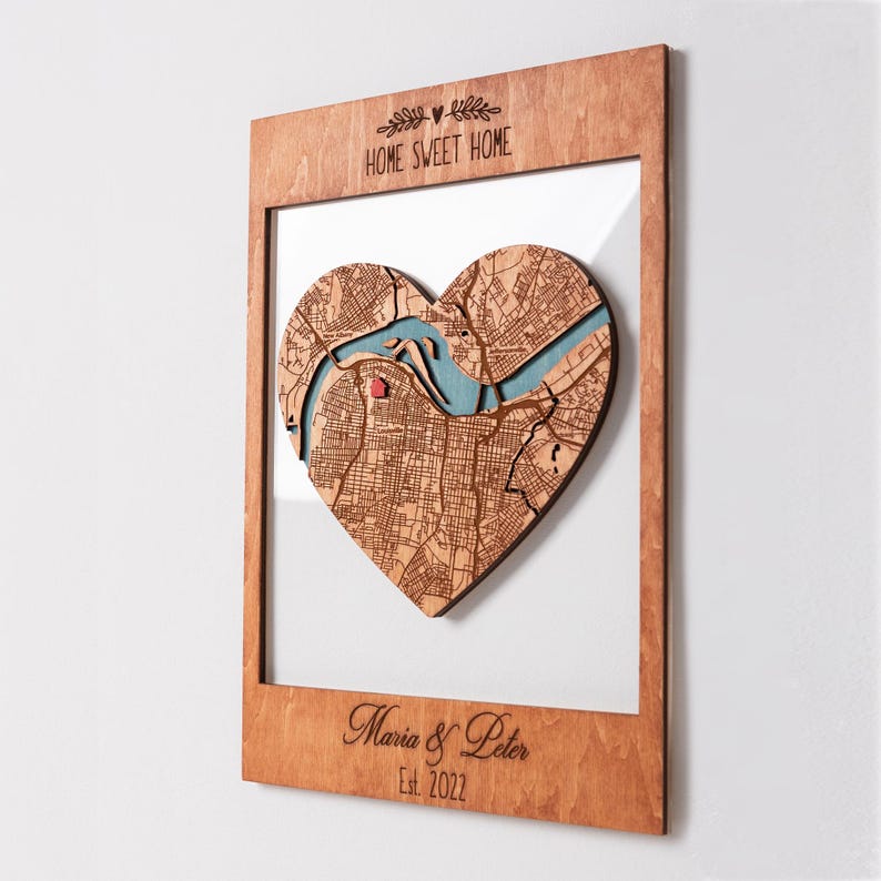 A framed heart-shaped map is shown from an angled view to highlight the depth and layered wood construction. This personalized wall art makes a thoughtful housewarming gift for a couple moving into a first home.