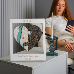 Personalized wooden heart map in a white frame displayed on a side table beside a candle, with a seated woman in the background. This scene presents the custom engagement and wedding gift as elegant shelf decor for a modern home.