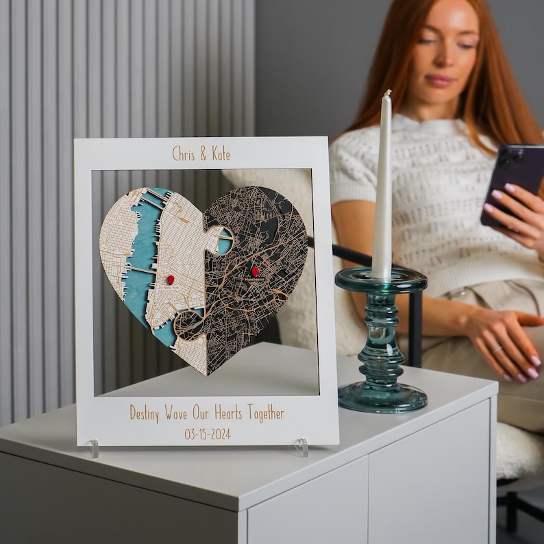 Personalized wooden heart map in a white frame displayed on a side table beside a candle, with a seated woman in the background. This scene presents the custom engagement and wedding gift as elegant shelf decor for a modern home.