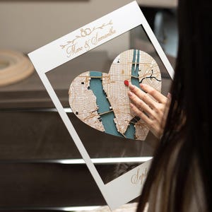 May include: White wooden frame with a heart-shaped map inside. The map is a 3D wood carving of a city with a red dot marking a location. The frame has the text "Marco & Samantha" and "Sept" written on it.