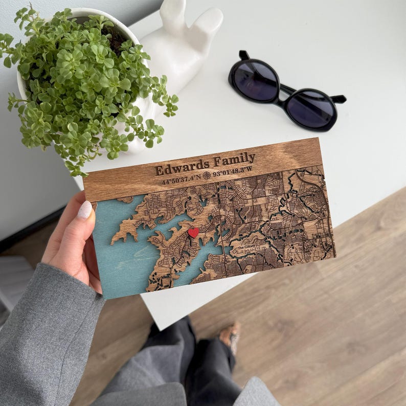 A hand holds a personalized wooden location map key holder above a desk with sunglasses and a plant nearby. This image shows the custom map detail and warm handmade finish before wall display.