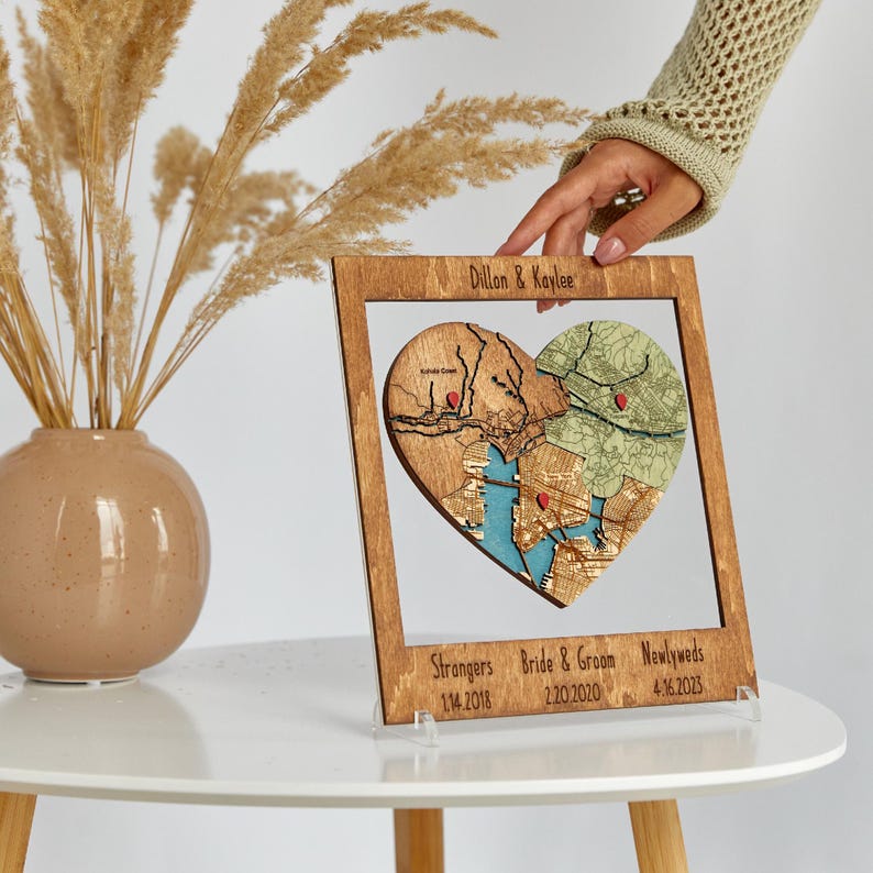 A hand adjusts the framed heart puzzle map while it stands on a round table beside a vase. This image highlights the piece as a decorative personalized keepsake for commemorating three relationship milestones.