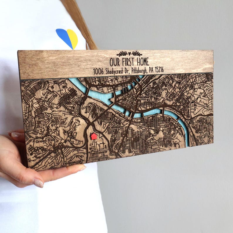 May include: A wooden map of a city with a light blue river and a red house marker. The text "OUR FIRST HOME" and address "1006 Shadycrest Dr, Pittsburgh, PA 15216" are engraved on the top. The map is held by a person.