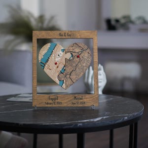 Personalized wooden heart map displayed on a dark round table with acrylic stands, showing two connected locations and engaged and married dates below. The scene presents the custom couple keepsake as romantic shelf decor with a polished, intimate look.