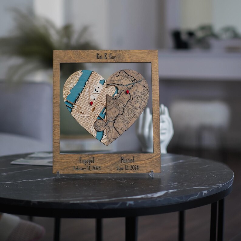 Personalized wooden heart map displayed on a dark round table with acrylic stands, showing two connected locations and engaged and married dates below. The scene presents the custom couple keepsake as romantic shelf decor with a polished, intimate look.